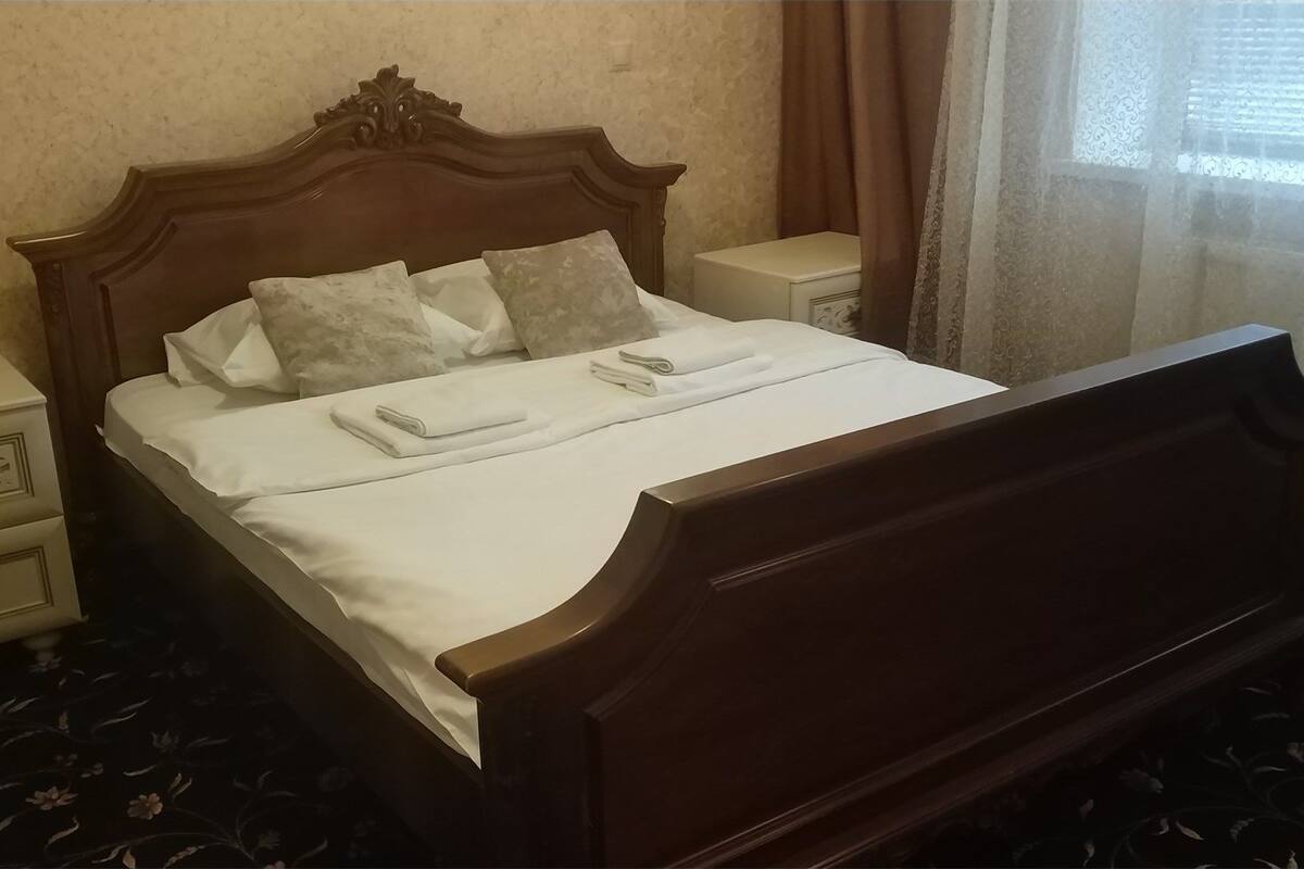 Un lit bien fait, avec une tête de lit en bois, du linge de lit blanc et deux oreillers. Il y a deux tables de chevet avec des lampes, une de chaque côté du lit. La pièce est dotée d'une moquette à motifs floraux et d'une fenêtre avec des rideaux.