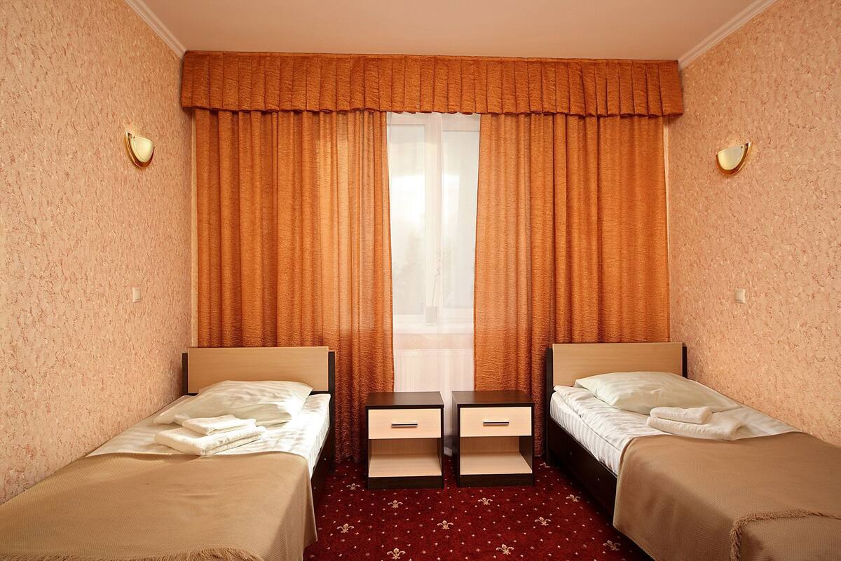 Une chambre d’hôtel avec deux lits simples, une fenêtre avec des rideaux, une moquette rouge et un luminaire fixé au mur.