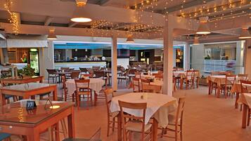 Een ruime restaurantruimte met meerdere eettafels en stoelen, sfeerverlichting en een bar op de achtergrond.