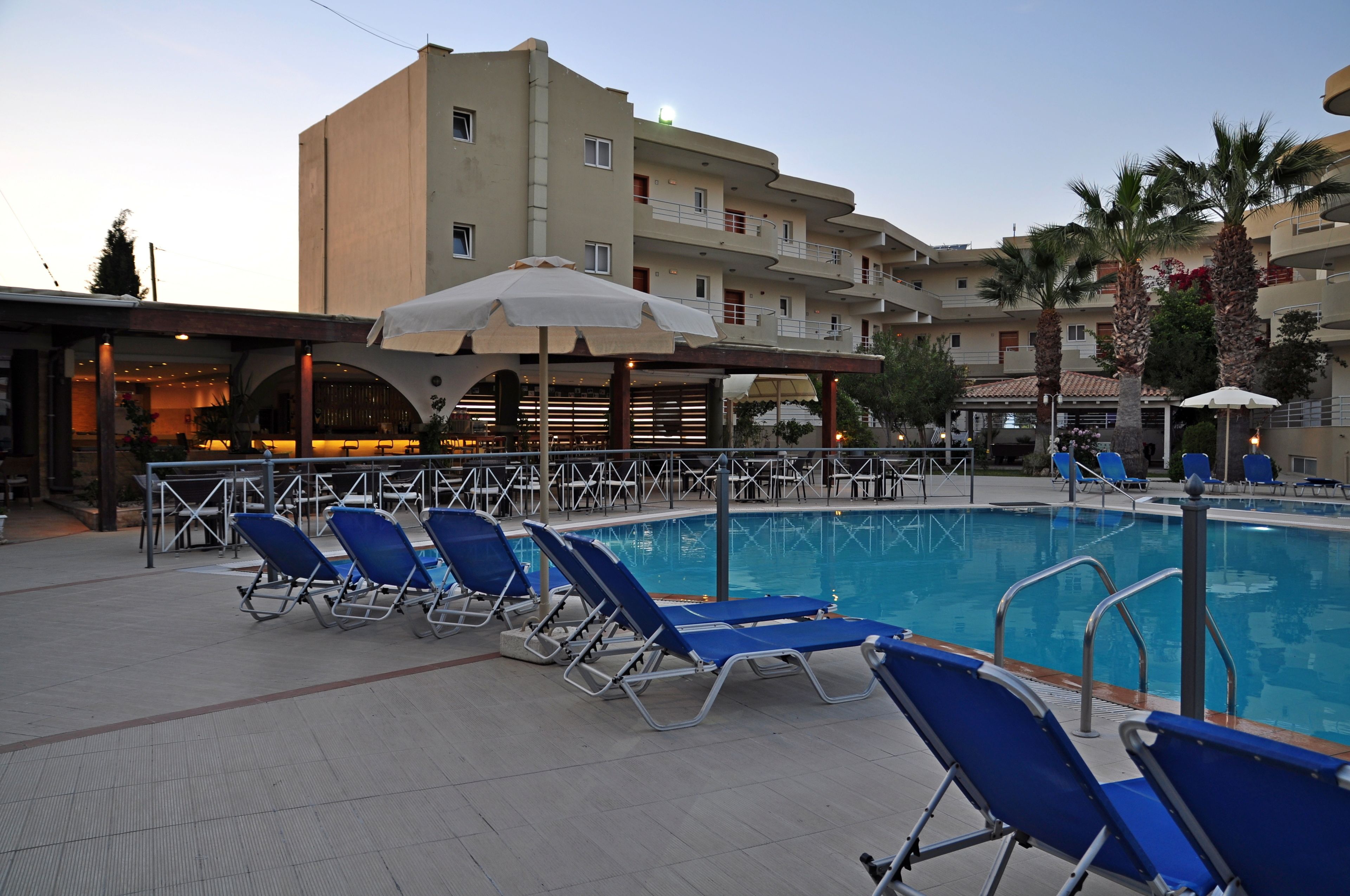 A swimming pool with lounge chairs and umbrellas, surrounded by a building with balconies and palm trees.