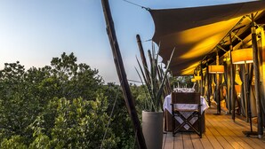 Un espace de restauration en plein air, avec une table dressée pour deux personnes, entouré d’une végétation luxuriante et protégé par une pergola qui offre de l’ombre.