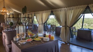 Un espace repas aménagé avec un buffet proposant divers plats et boissons, situé dans un hébergement sous tente offrant une vue sur l’extérieur.