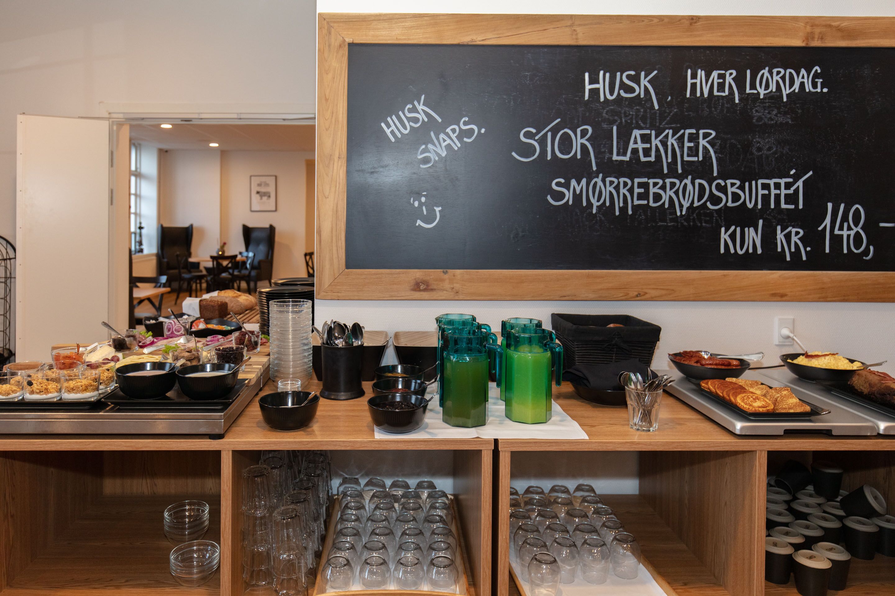 Un espace buffet proposant divers plats et boissons, un menu écrit à la craie et un coin salon à l’arrière-plan.