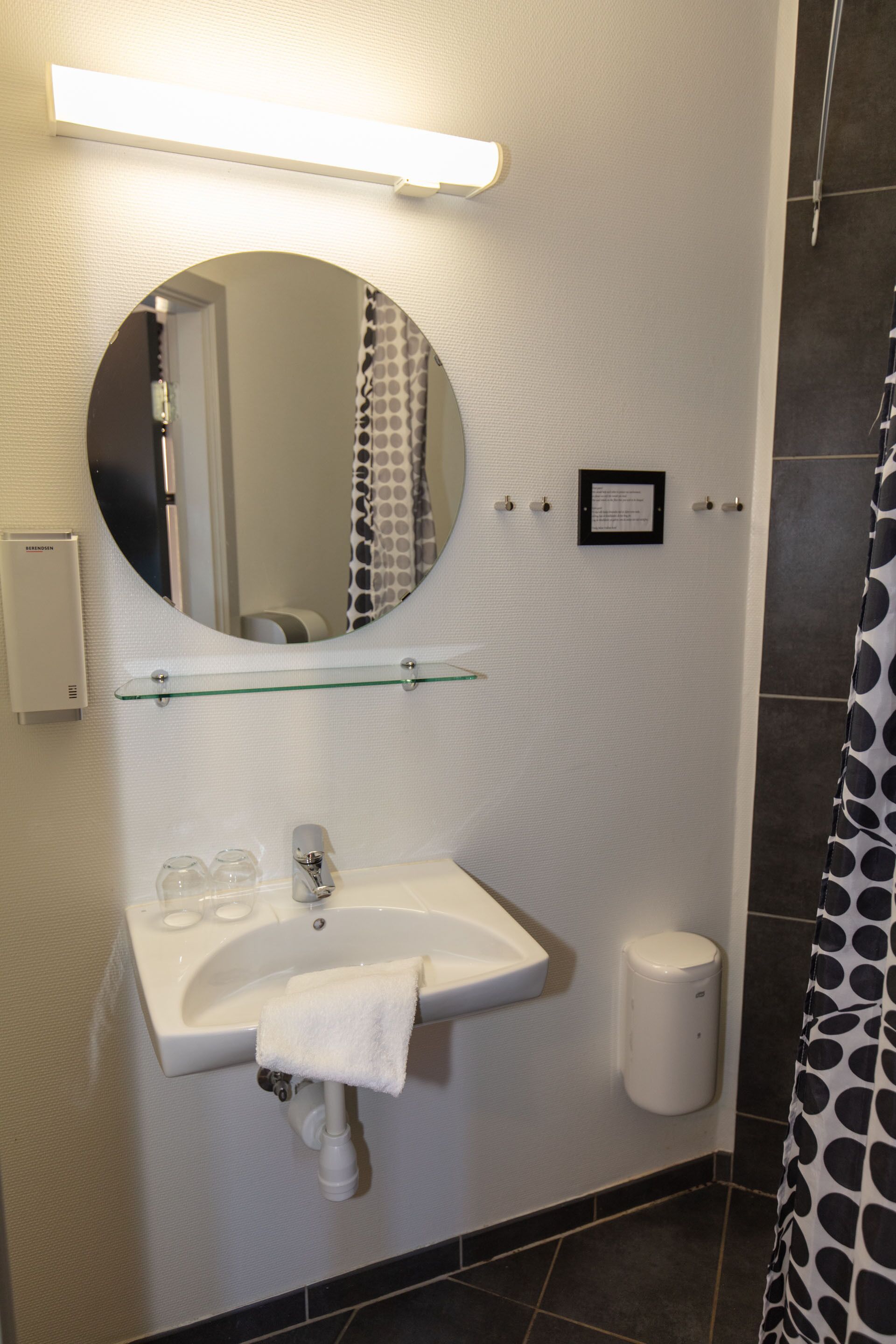 A bathroom with a round mirror, a glass shelf, a white sink, and a towel hanging on the wall.