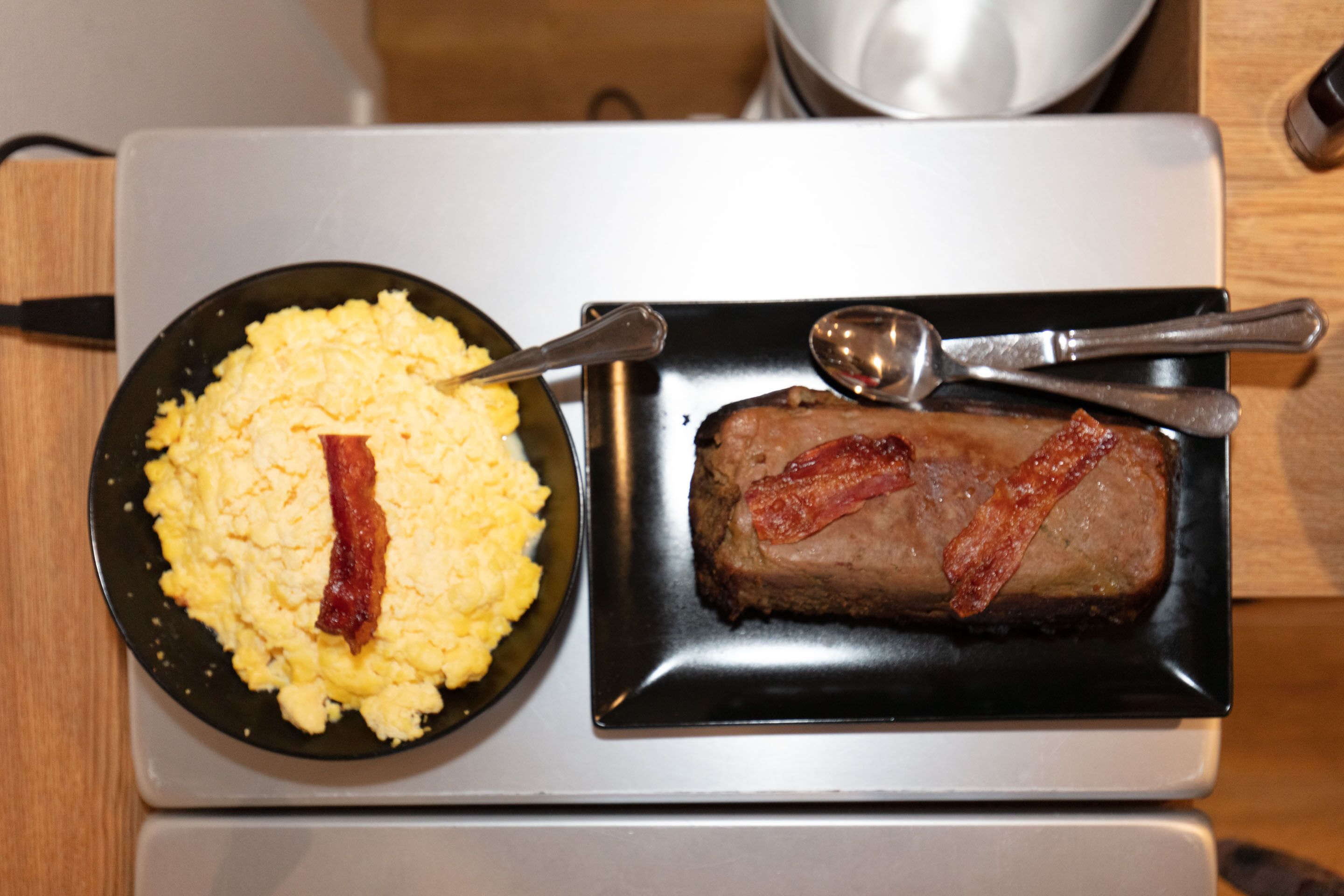 Un plato con huevos revueltos y tocino, además de un filete también con tocino, sobre un plato negro.