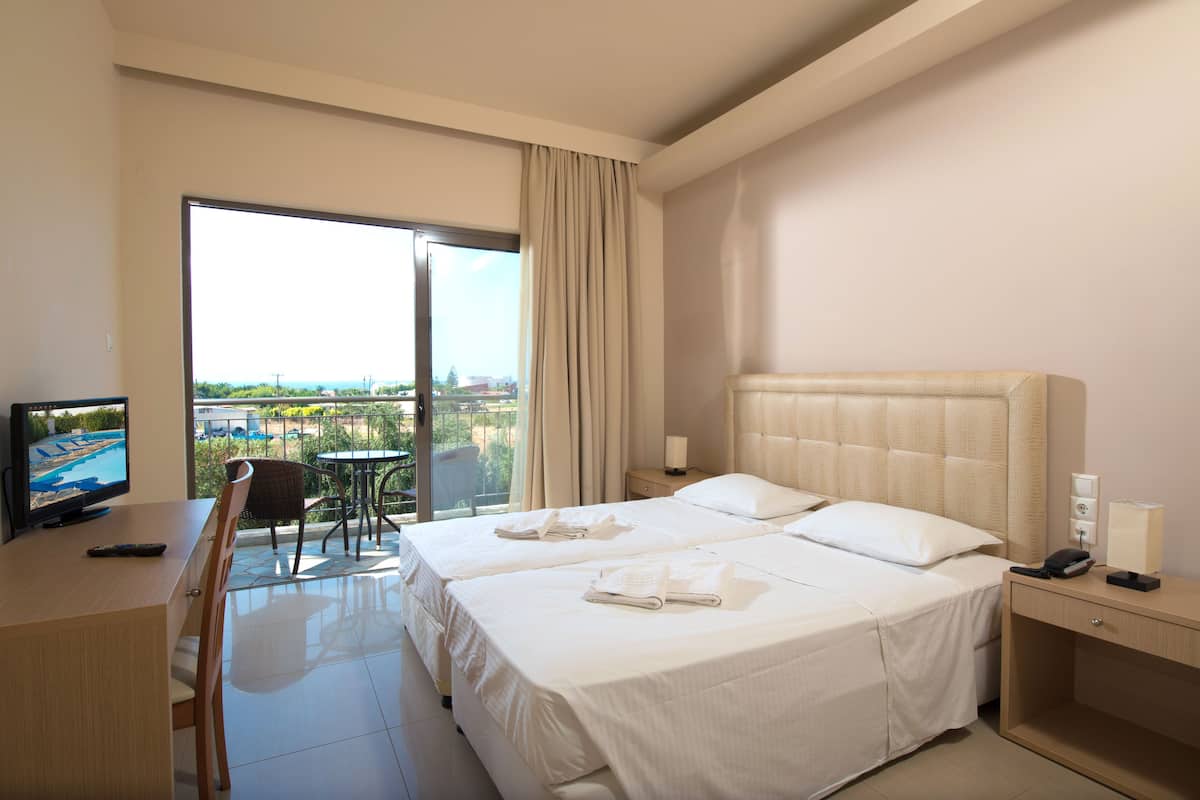 Habitación de hotel con dos camas, televisión, escritorio y balcón con vistas.