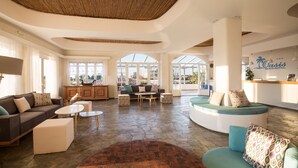 Un hall d’entrée spacieux doté d’un sol en pierre, d’un comptoir de réception, d’un espace salon avec des canapés et une table basse, et de grandes fenêtres offrant une vue sur l’extérieur.