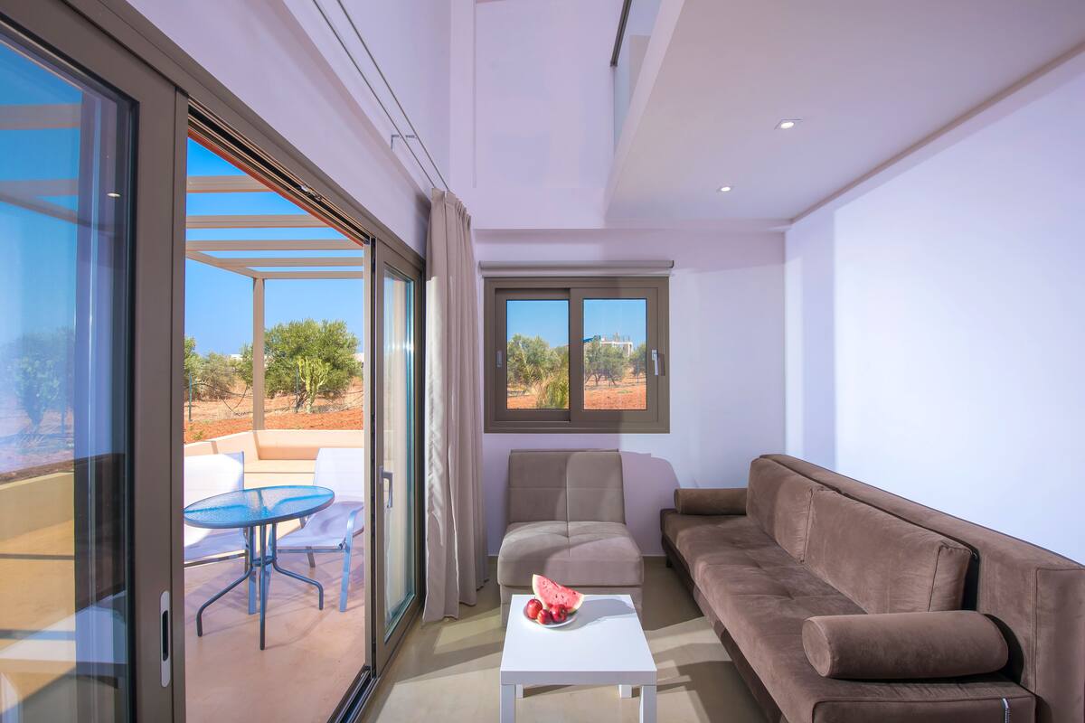 Una sala de estar moderna con un ventanal que da a un espacio al aire libre, un sofá marrón, una mesa blanca con fruta y vistas a árboles y edificios.