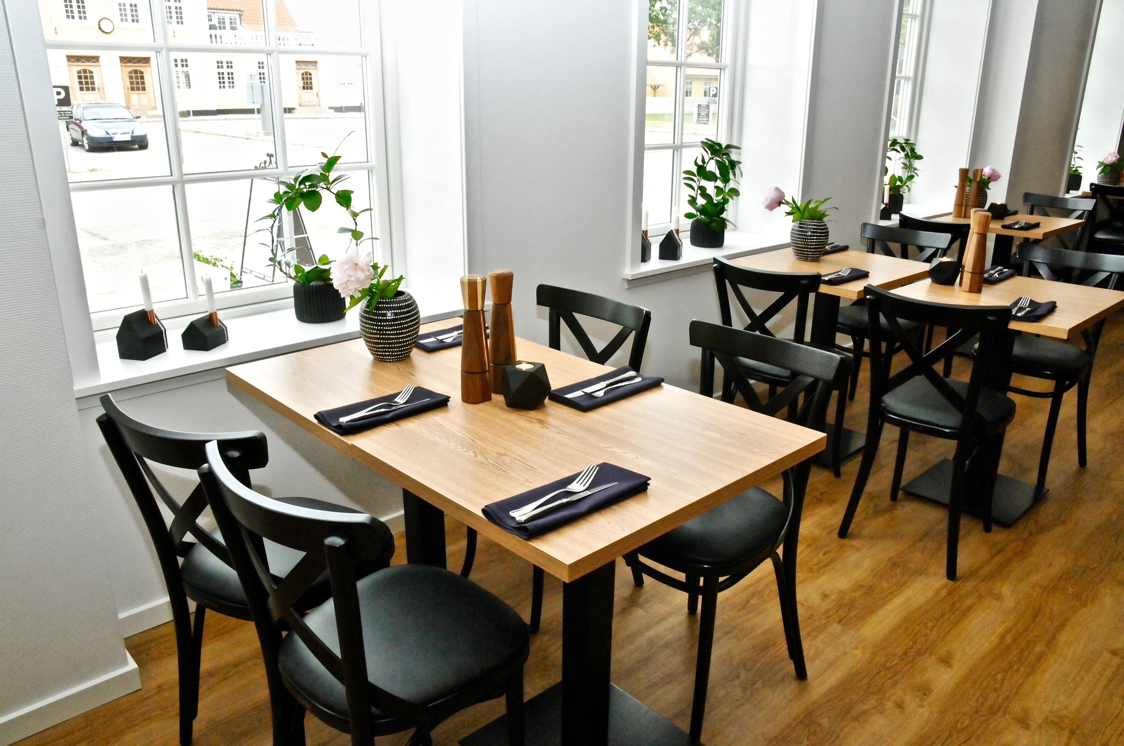 Un restaurante con mesas de madera y sillas negras, plantas en macetas y vistas de la calle a través de amplios ventanales.
