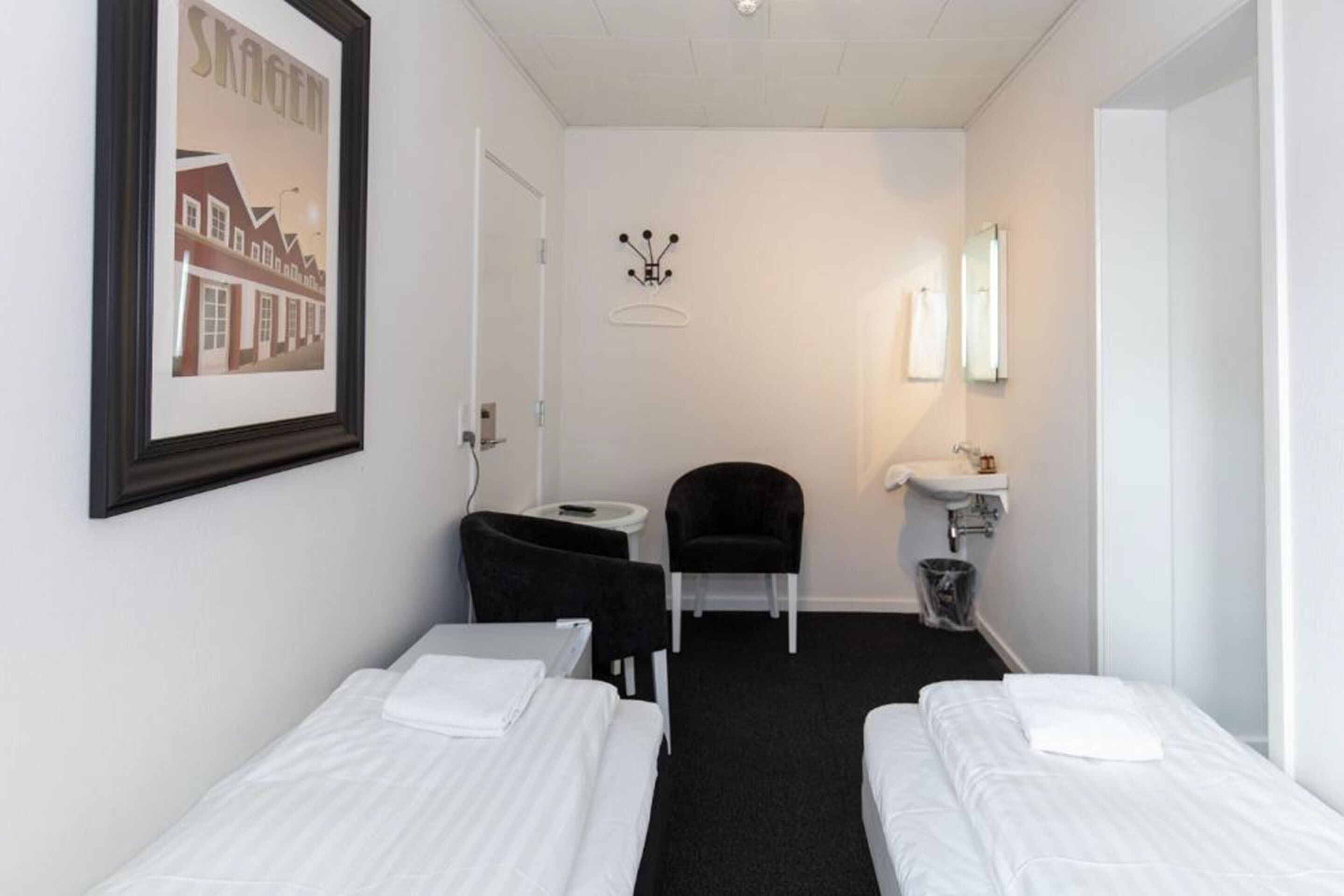 A small hotel room with two single beds, a desk, a chair, a small table, a mirror, and a framed picture on the wall.