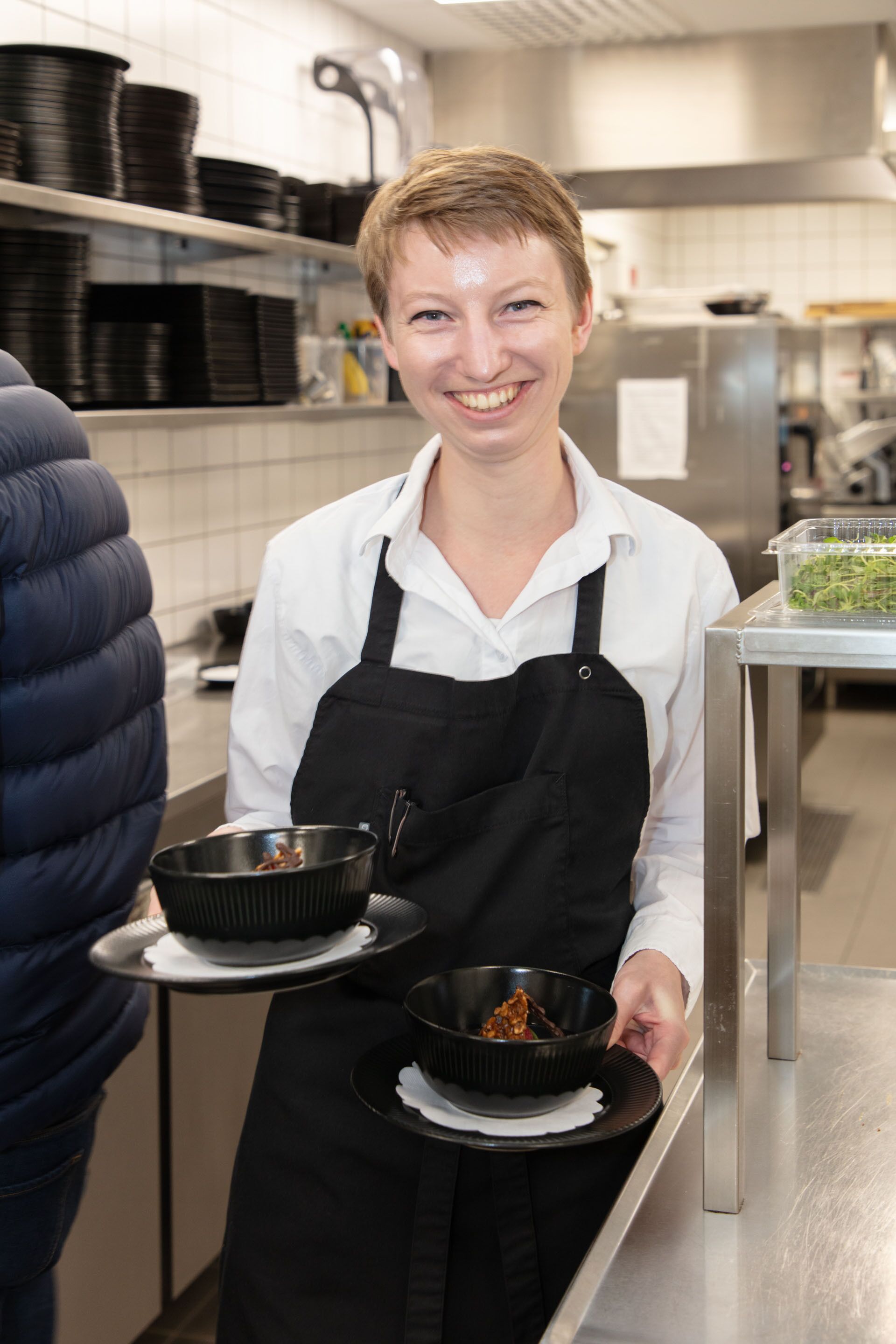 A person in a white shirt and black apron holding two black bowls on a tray.