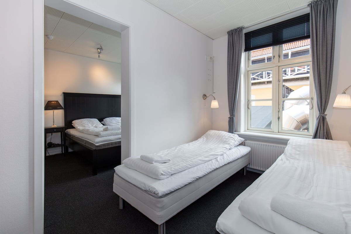 Habitación de hotel con una cama individual, una mesita de noche pequeña con lámpara, una ventana con cortinas y vistas al balcón exterior.