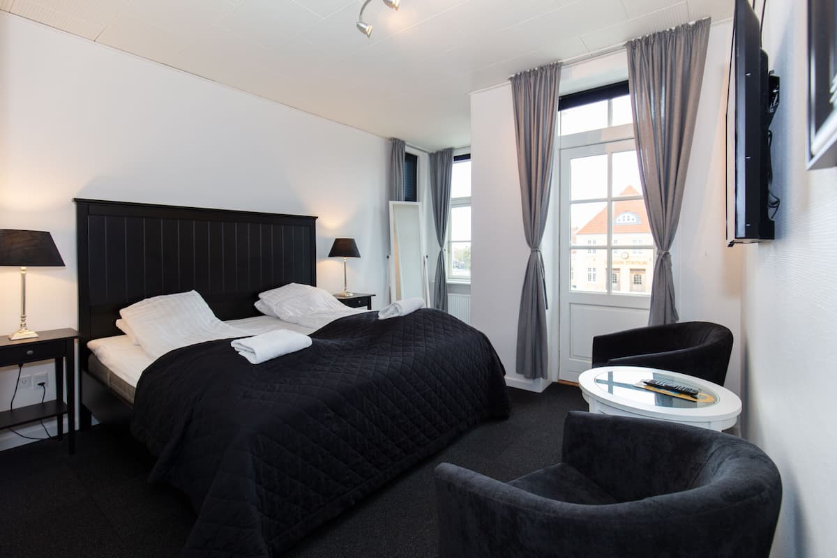 Una habitación de hotel con una cama grande, una mesita de noche con lámpara, una silla, una pequeña mesa redonda con un control remoto y un televisor montado en la pared.