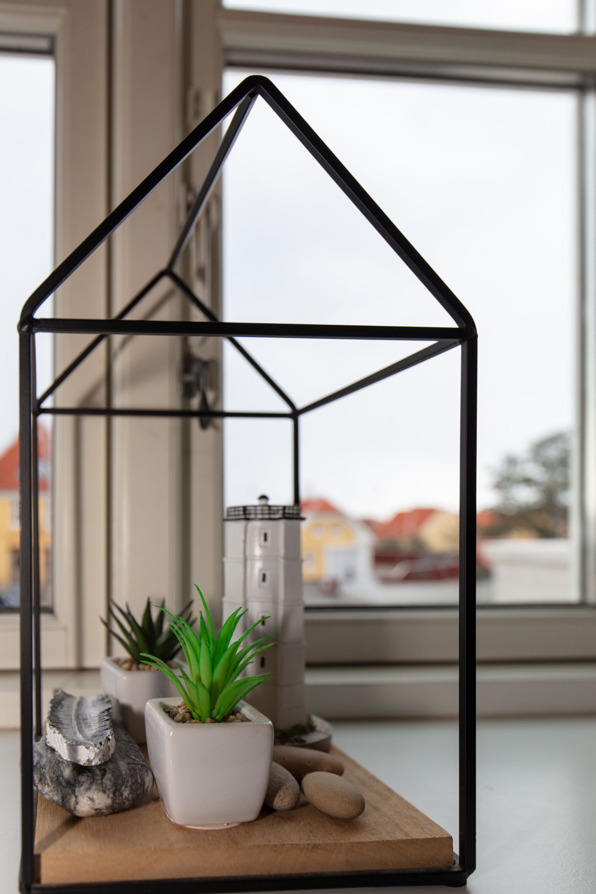 Un ambiente interior minimalista con una decoración en forma de casa geométrica, una planta en maceta y un objeto blanco pequeño sobre una superficie de madera.