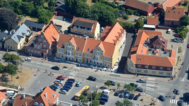 An aerial view of a town with red-roofed buildings, a central square, and a busy street with cars and pedestrians.