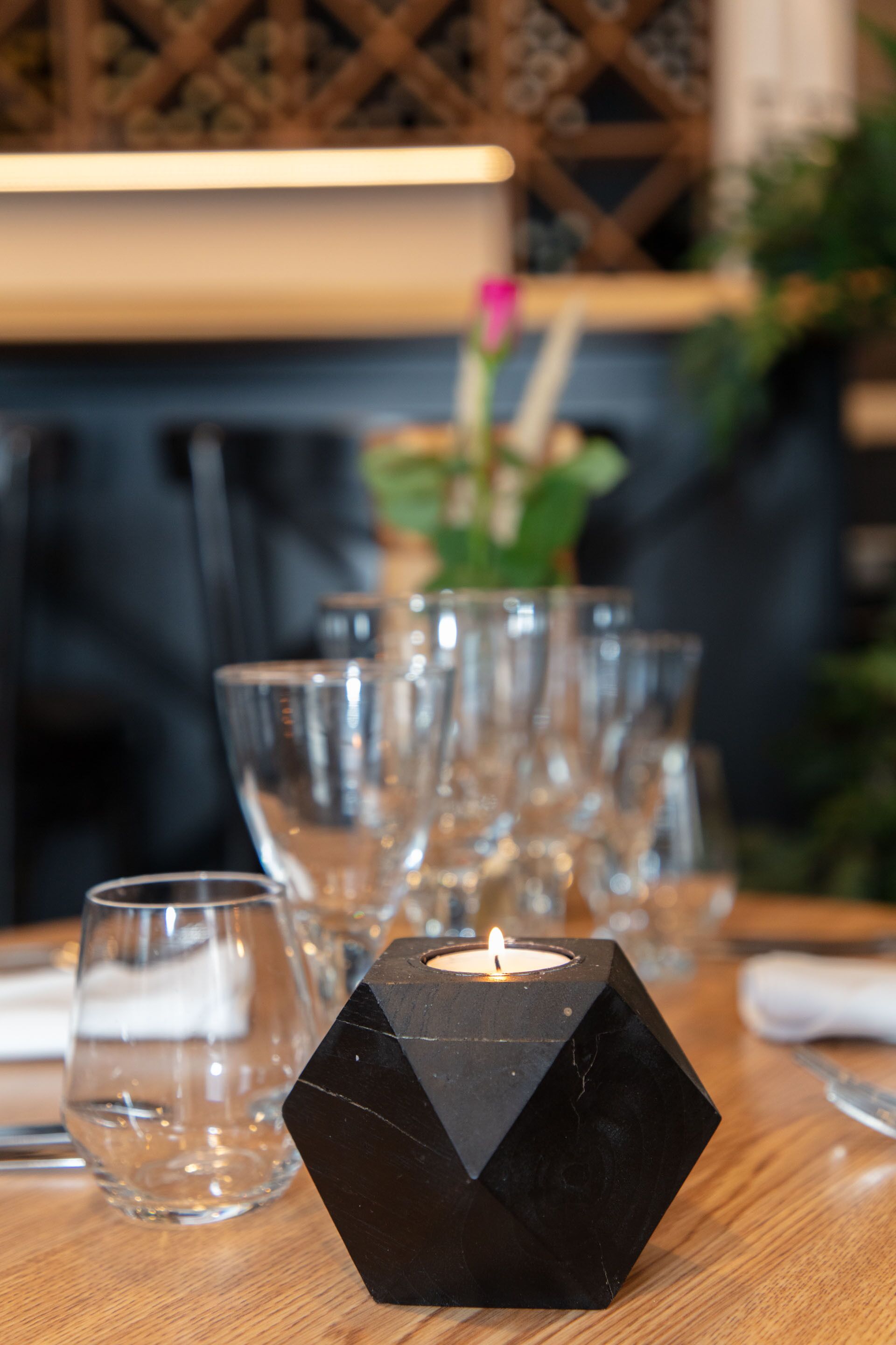 Un candelabro hexagonal con una vela encendida sobre una mesa de madera, rodeado de vasos transparentes y un florero con una flor rosa.