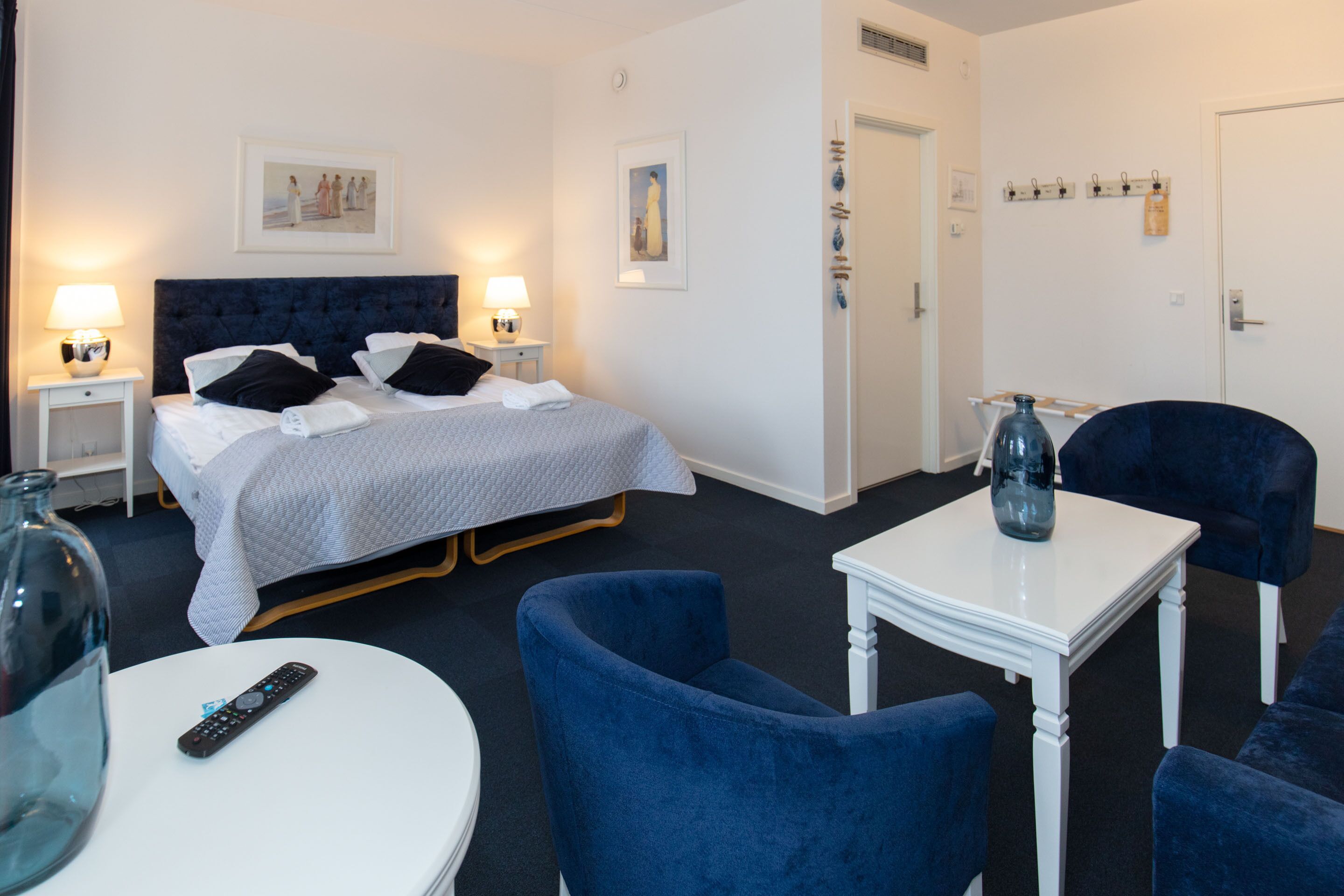 Habitación de hotel con una cama, dos mesitas de noche con lámparas, una mesa blanca con un jarrón y dos sillones azules.