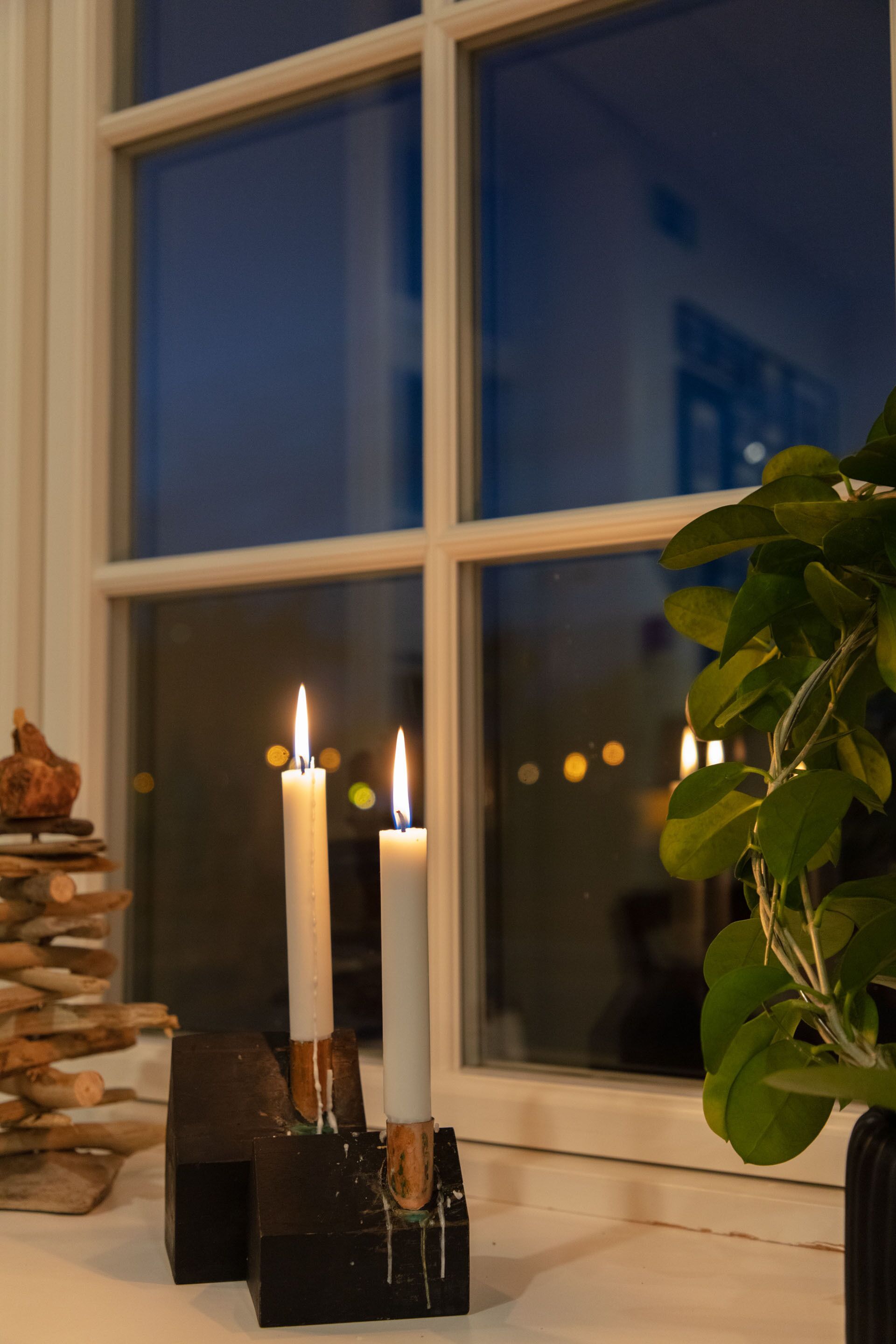 Uma janela com três velas acesas em suporte preto, uma planta à direita e uma pilha de lenha à esquerda.