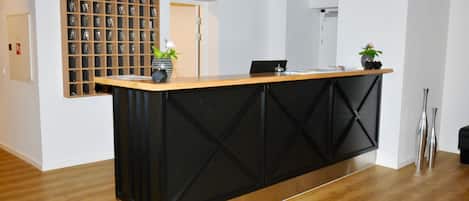 A reception desk with a black and wooden design, a wall-mounted display of glassware, and two potted plants.