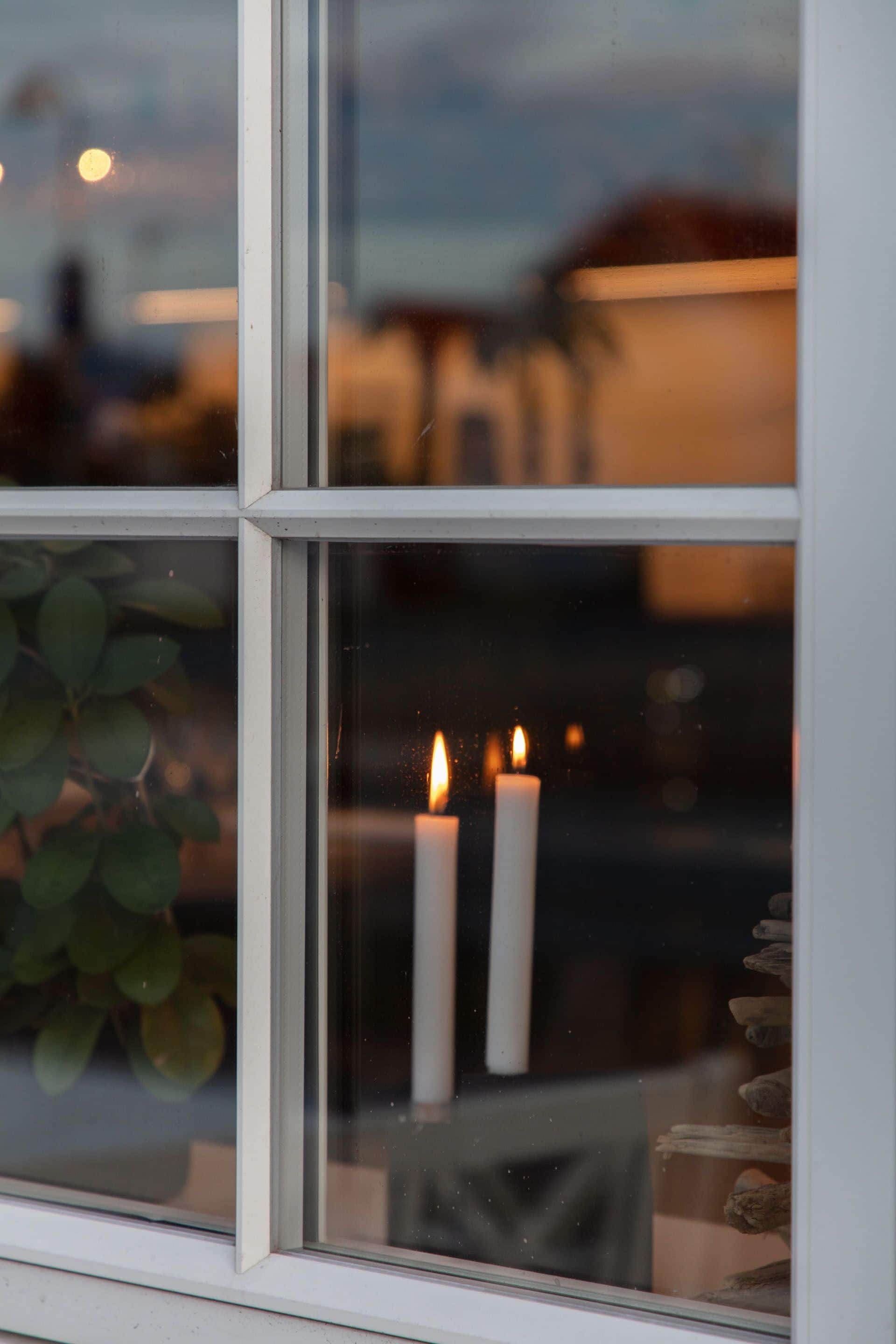 A window with two lit candles, a plant, and a view of a building outside.