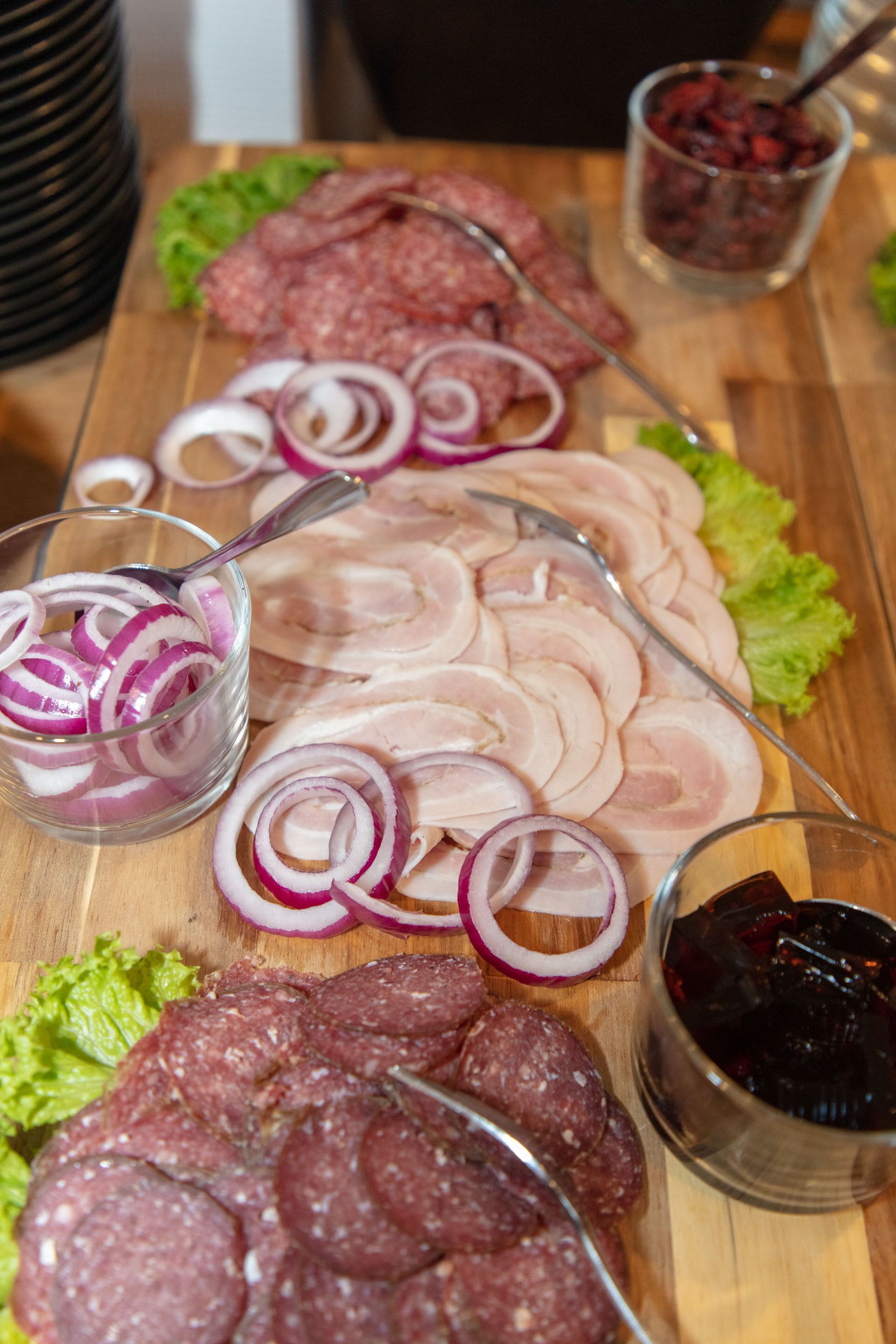 A wooden board with sliced meats, onions, and a glass of red condiment.