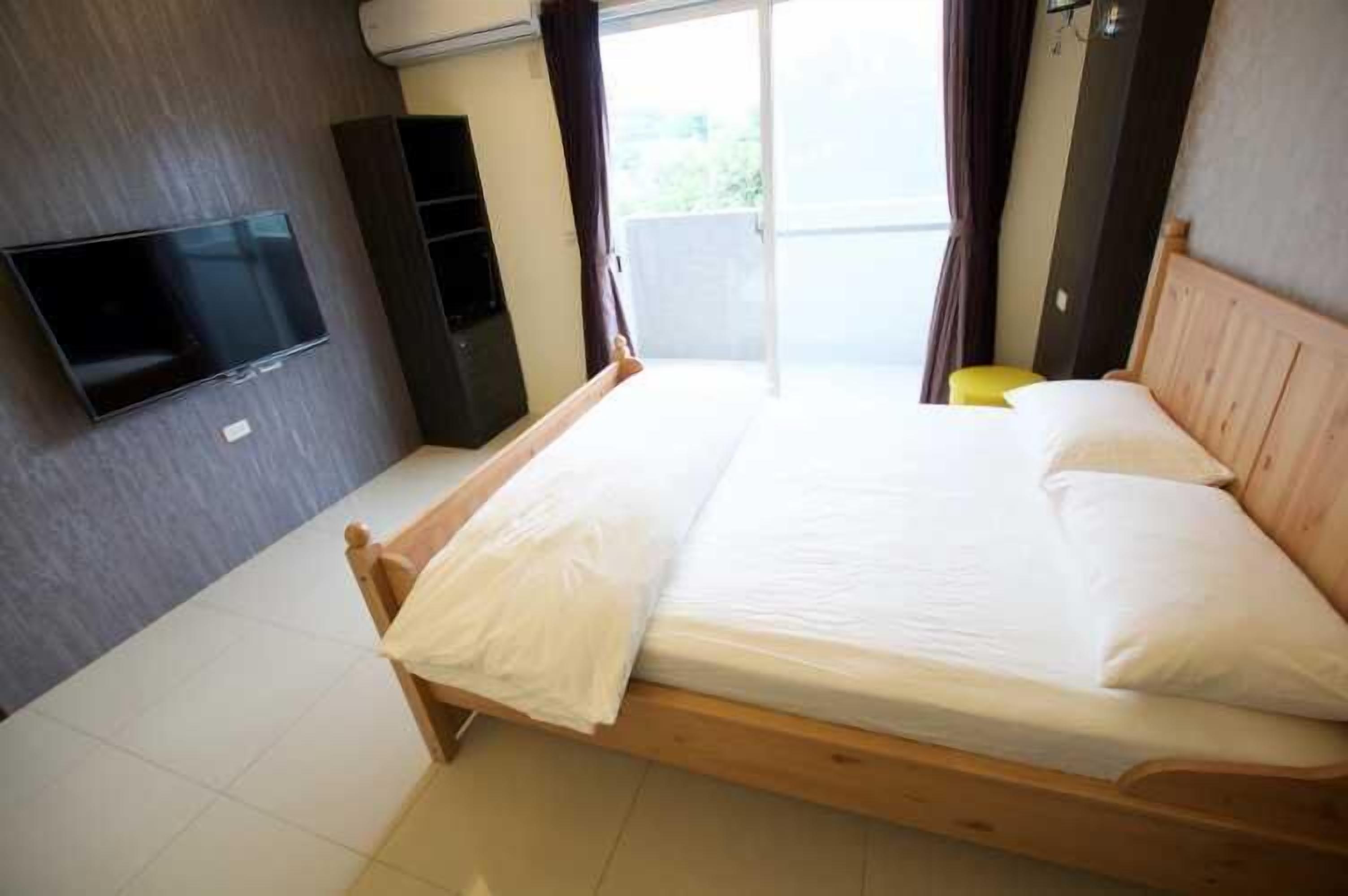A bedroom with a wooden bed, a flat-screen TV mounted on the wall, and a sliding glass door leading to a balcony.