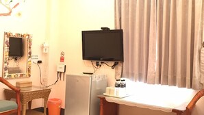 Una habitación pequeña con televisión, refrigerador, una mesa con una bandeja de objetos, una silla de mimbre y una ventana con cortinas.