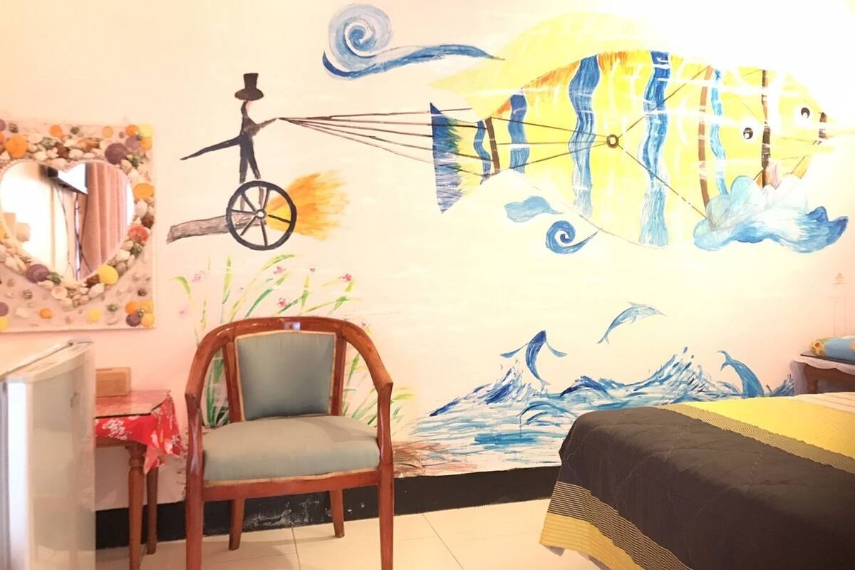 Una habitación con una cama, una silla, un escritorio y un mural que muestra una escena de circo y un pez.