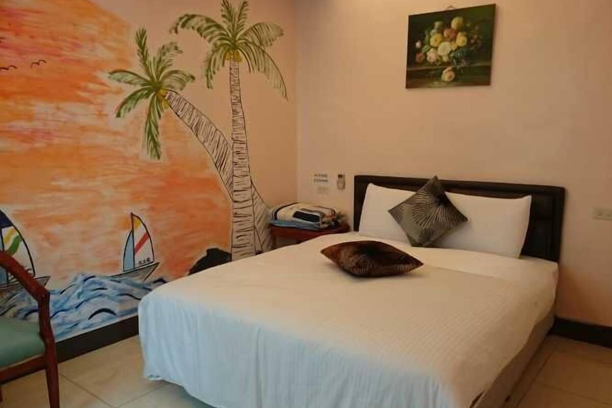 Un dormitorio con una cama, una mesita de noche, un cuadro de palmeras y un mural en la pared.