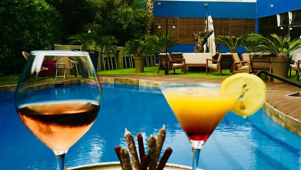 Een tafeltje bij het zwembad met twee drankjes, een bord eten en een zwembad op de achtergrond.