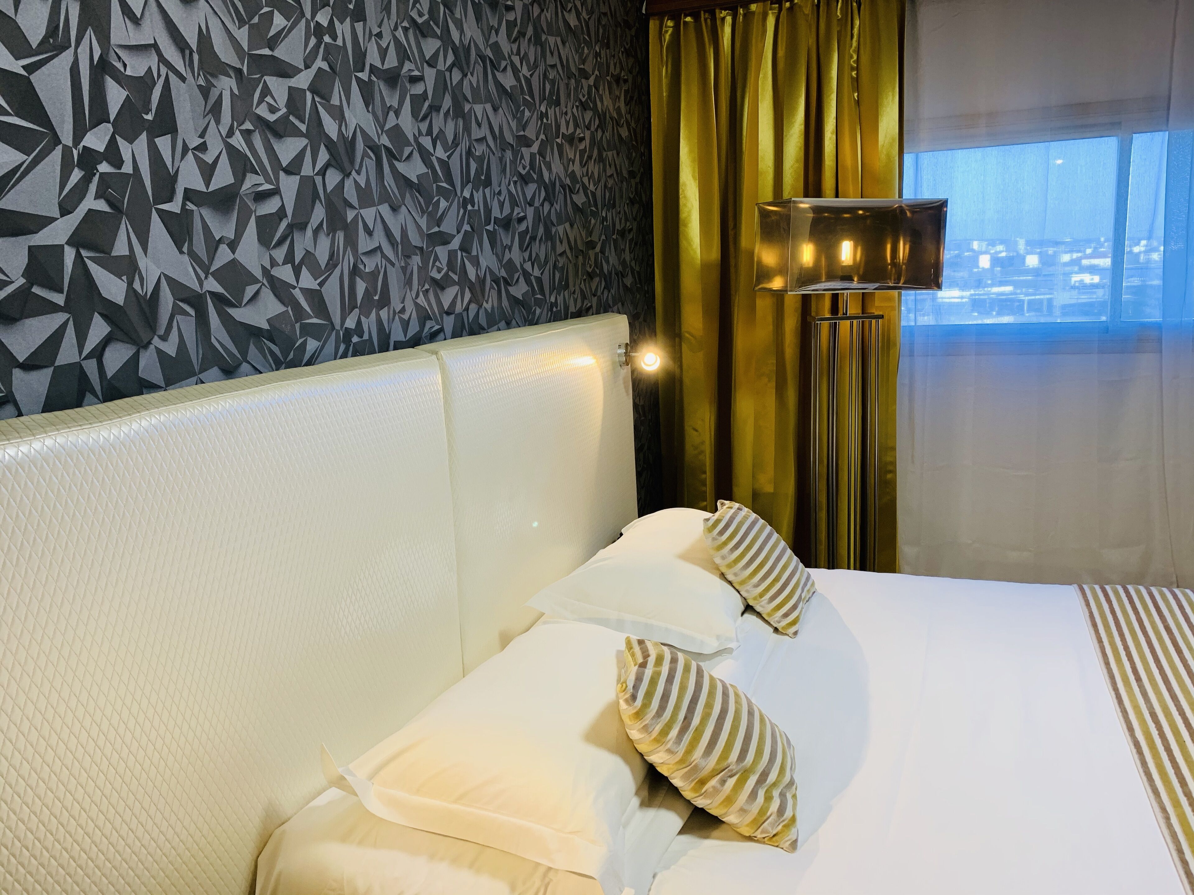 Una habitación de hotel con una pared de diseño geométrico, un ventanal con cortinas y una cama con almohadones a rayas.