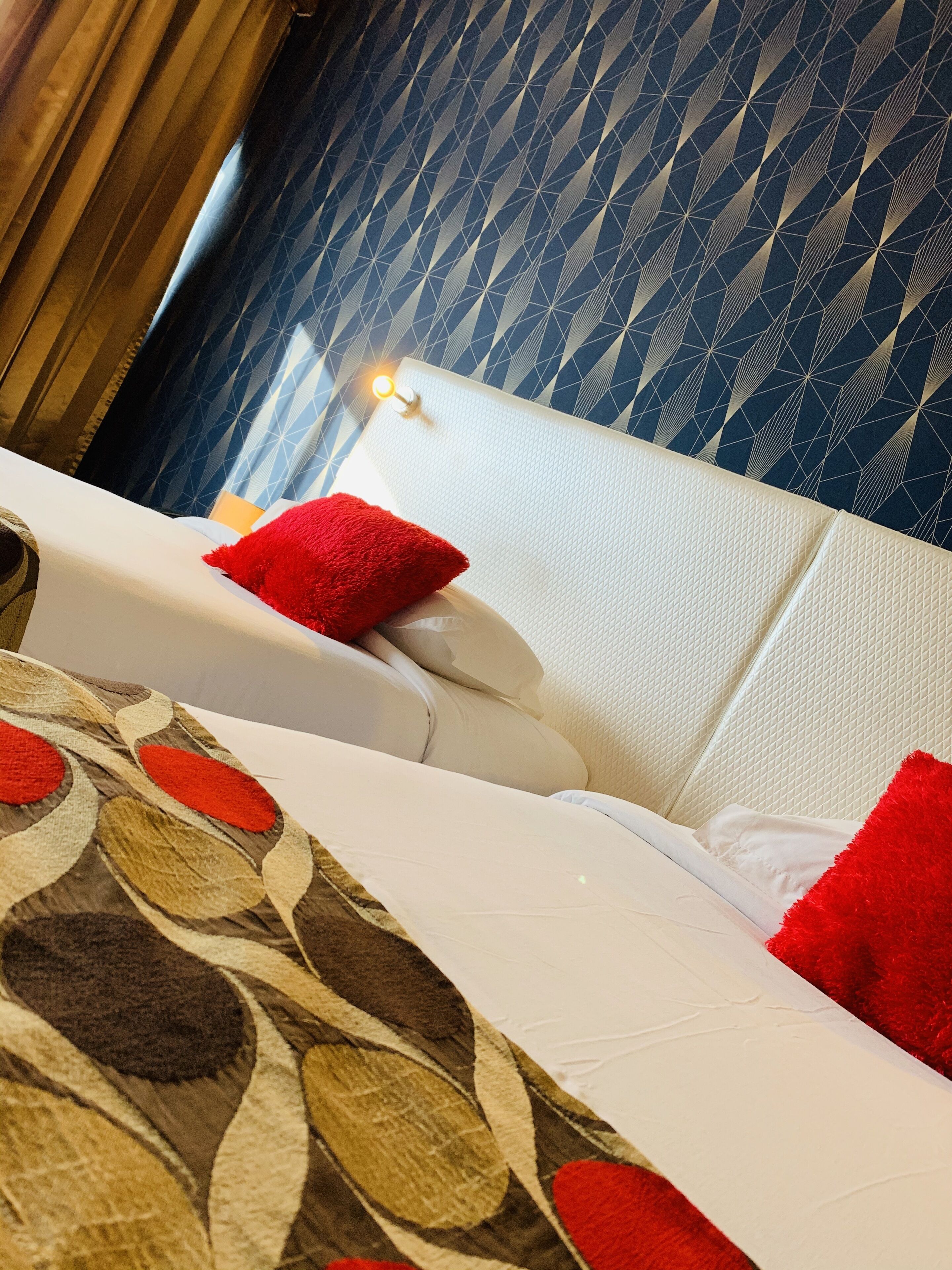 Una habitación de hotel con cama, almohadas rojas, un cabecero estampado y una ventana con cortinas.