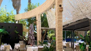 Een overdekte eethoek met tafels en stoelen, omgeven door groen, onder een pergola.