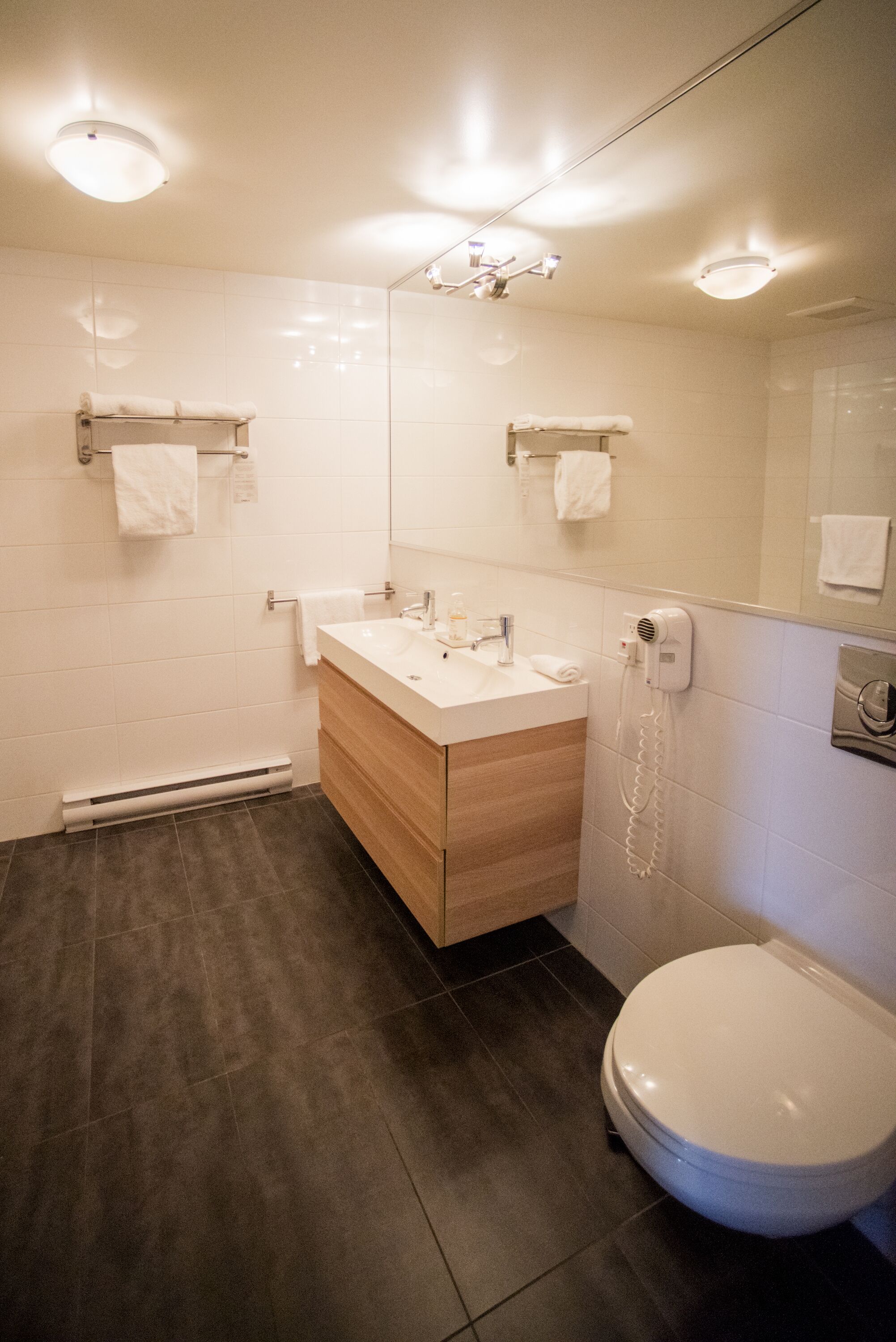 A modern bathroom with a white sink, wooden vanity, and a toilet. There are two towel racks on the wall and a hairdryer on the floor.
