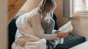 A cozy room with wooden beams, a window with curtains, a lamp, and a person reading a book. - Hotel Horizon (Sutton)