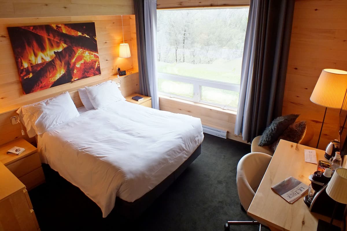 Une chambre d’hôtel avec un grand lit, un bureau avec une chaise, une fenêtre avec des rideaux, une lampe fixée au mur et un tableau représentant un feu.