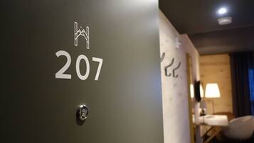 Une porte de chambre d’hôtel portant le numéro 207, un trou de serrure et une vue partielle sur un lit et une table de chevet avec une lampe.