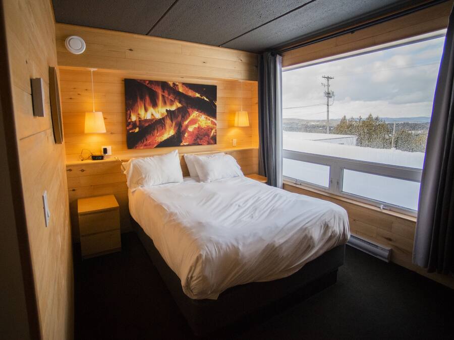 A hotel room with a bed, bedside lamps, a painting on the wall, and a window with a view of a snowy landscape.