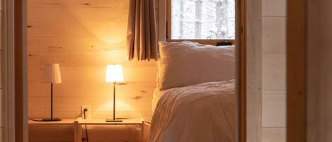 Un dormitorio acogedor con piso de madera, una cama con ropa de cama blanca, una mesita de noche con una lámpara y una ventana con cortinas.