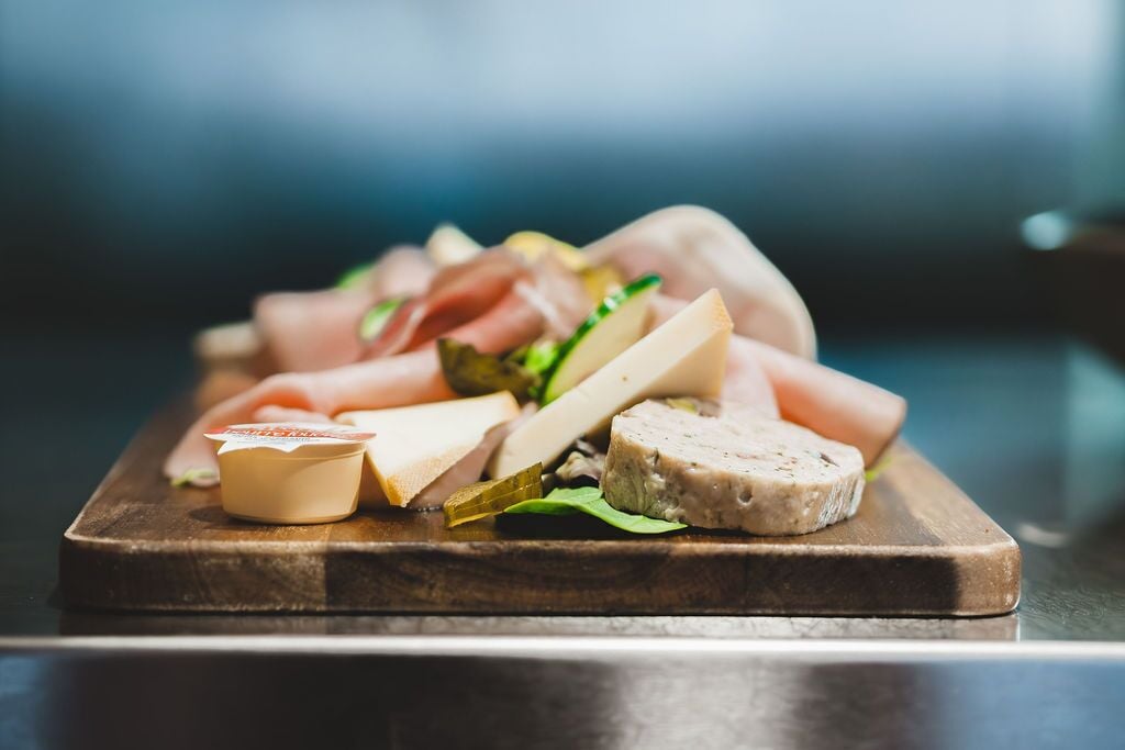 A wooden board with various cheeses, cured meats, and a small container of butter.