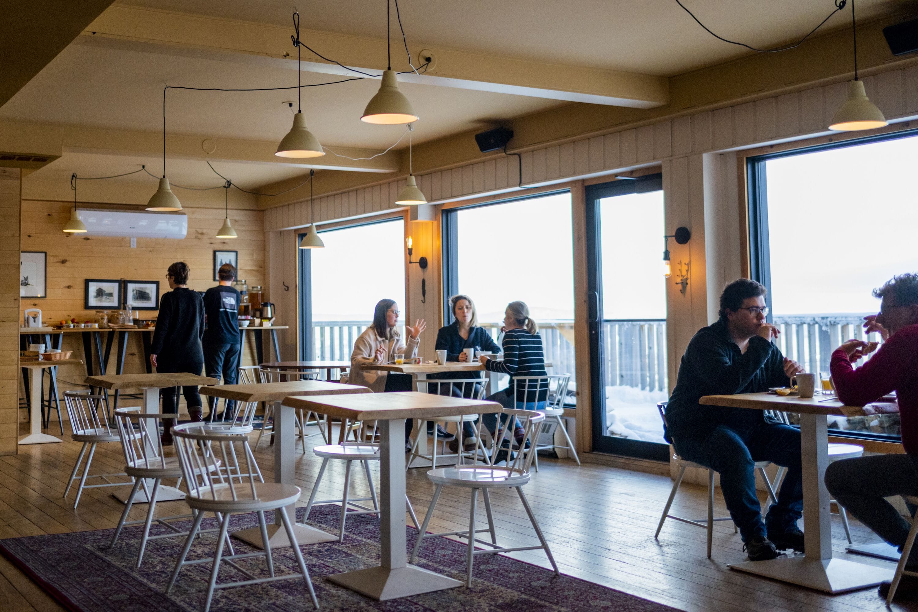 Un café con mesas y sillas de madera, ventanas amplias y lámparas colgantes.