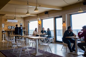 Um café com mesas e cadeiras de madeira, janelas amplas e luminárias pendentes.