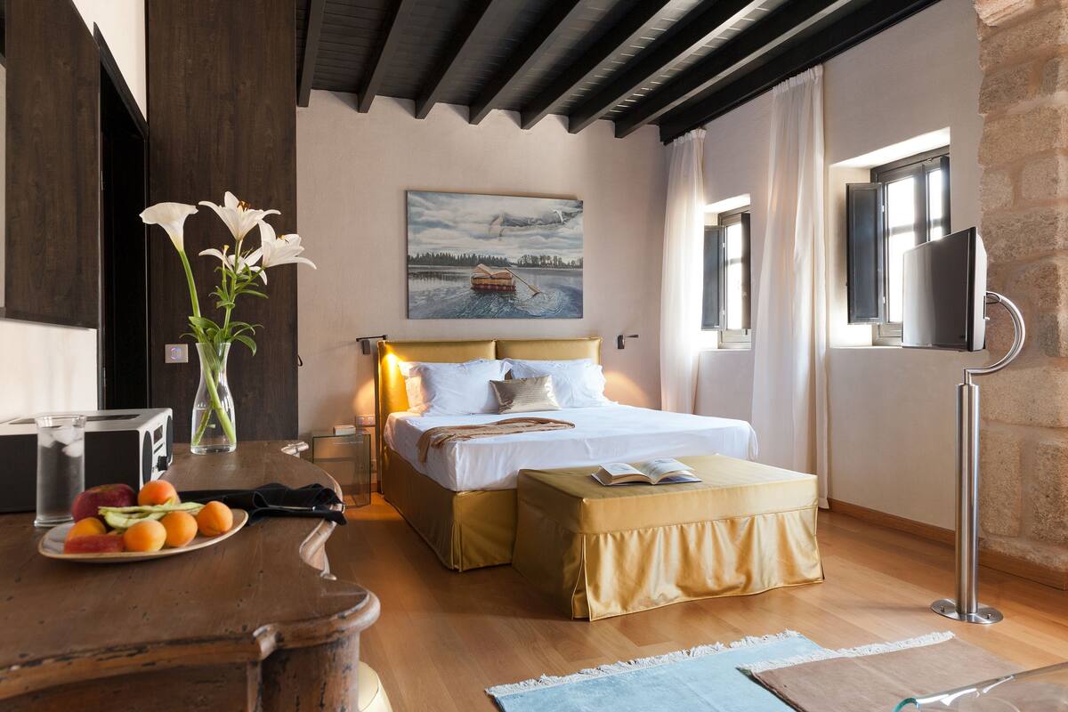 A bedroom with a bed, bedside table, a painting, a window, and a TV.