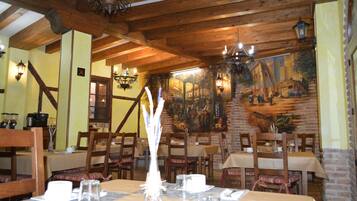A restaurant with wooden beams, brick walls, and a mural. Tables are set with white tablecloths and candles.