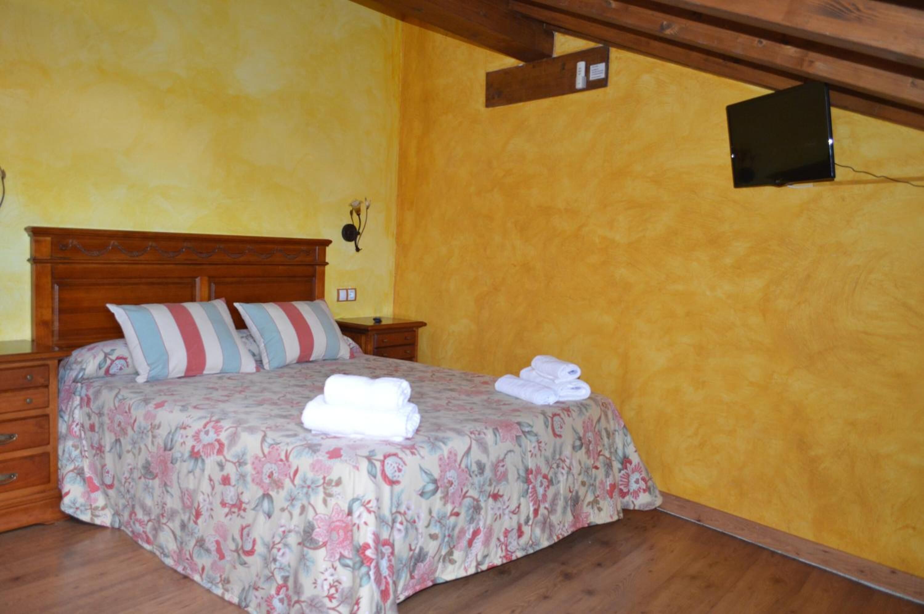 Un dormitorio con cama de madera, colcha estampada, almohadas a rayas, una mesita de noche y un televisor montado en la pared.