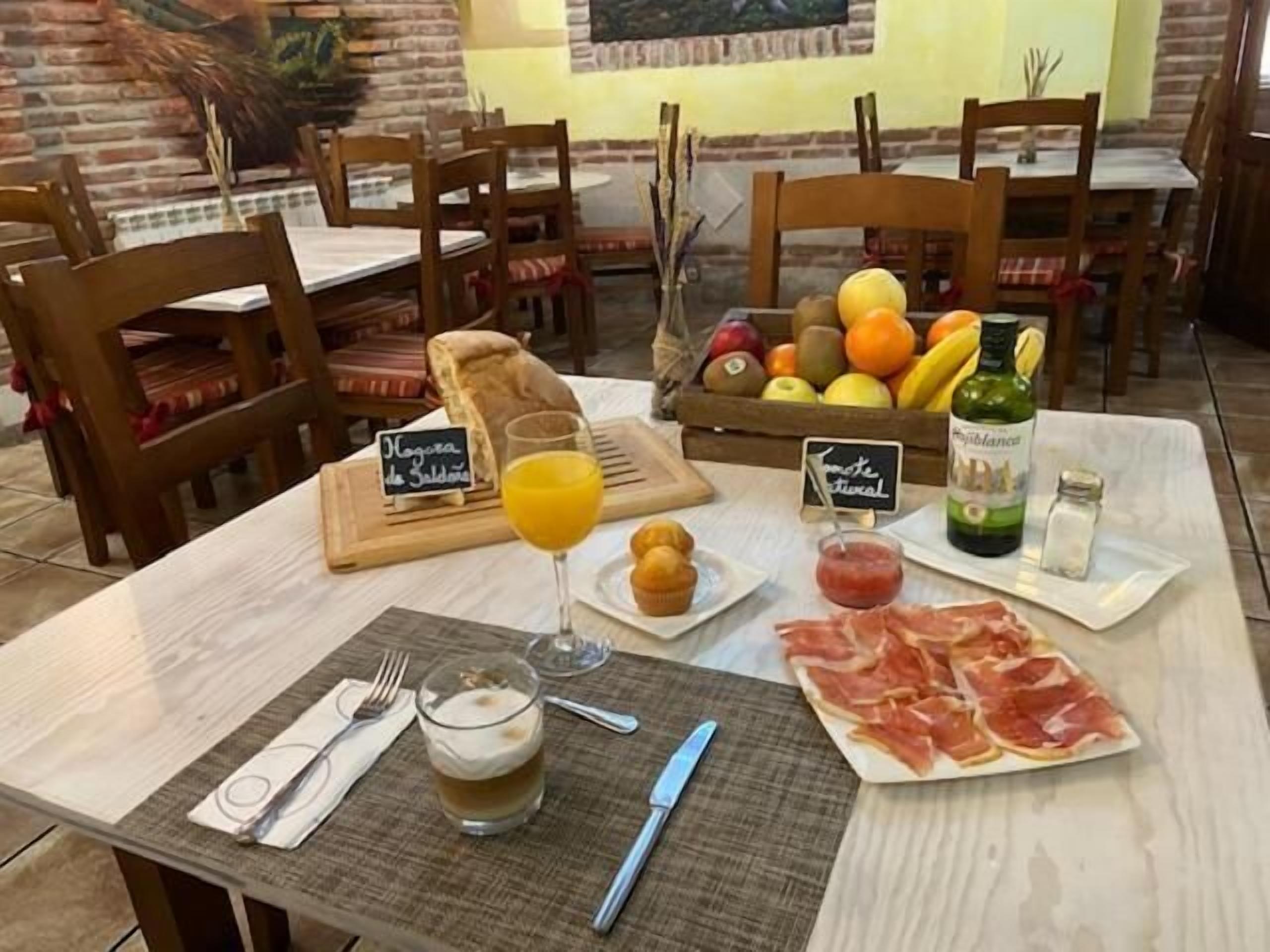 A breakfast setting with a table set for two, featuring bread, fruit, a glass of orange juice, a plate of prosciutto, a bottle of olive oil, and a glass of milk.