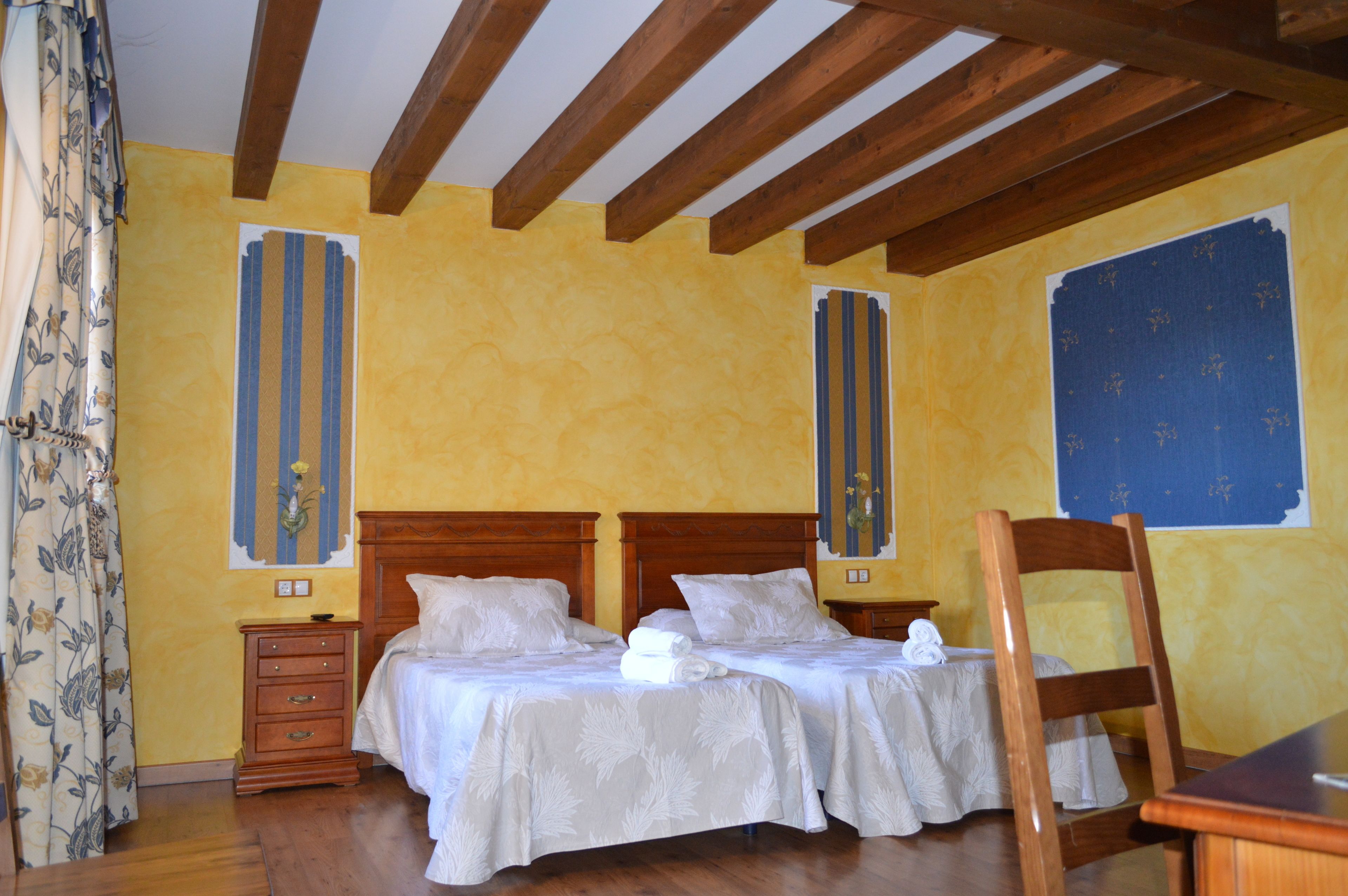 Una habitación con dos camas, cabeceros de madera, un aparador de madera y un ventanal con cortina azul estampada.