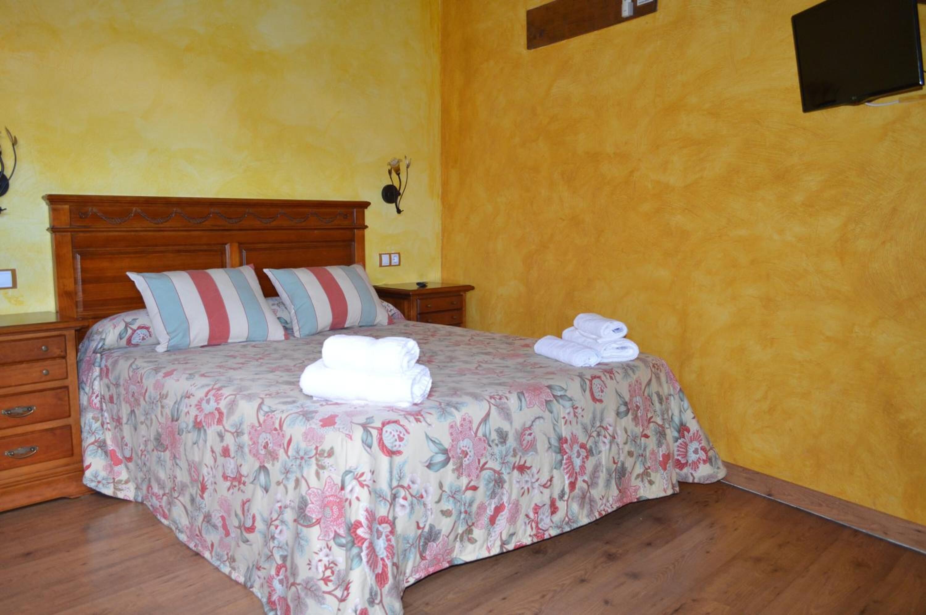 A bedroom with a floral bedspread, two pillows, a wooden headboard, a nightstand, and a wall-mounted clock.