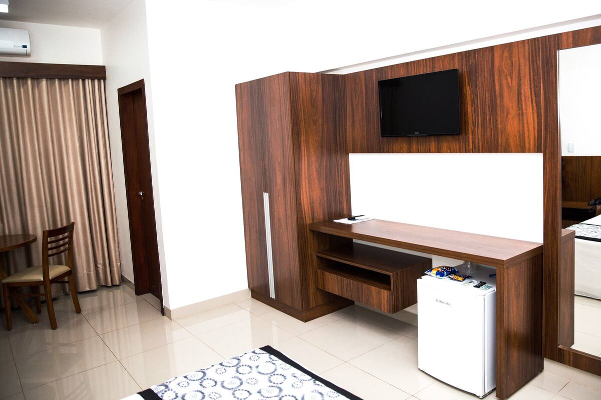 A compact hotel room with a wooden desk, a flat-screen TV, a minibar, and a bed with a patterned bedspread.