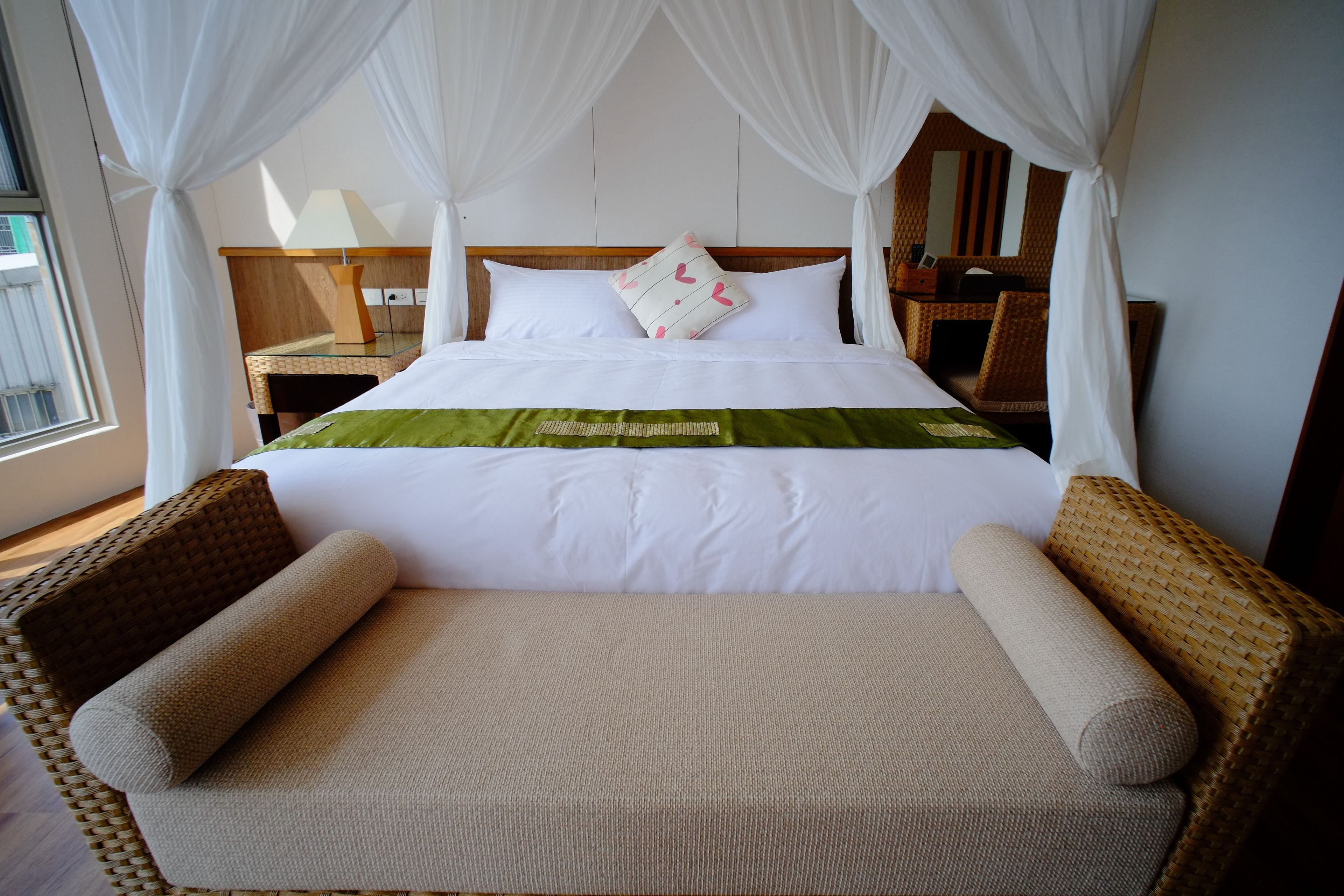 A neatly made bed with white linens and a green accent, flanked by wicker chairs.